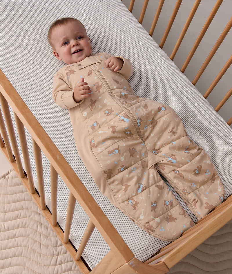 A happy baby lying down inside a wooden cot on a subtle striped mattress. The baby is wearing a light brown 2.5 TOG Sleep Suit Bag with long beige sleeves, a front zipper, and a gentle pattern of small brown animals and light blue accents.