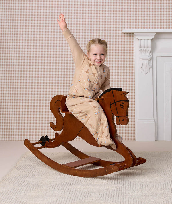 Cheerful preschooler wearing a beige patterned sleep suit bag, riding a wooden rocking horse with one arm raised in the air.