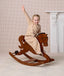 Cheerful preschooler wearing a beige patterned sleep suit bag, riding a wooden rocking horse with one arm raised in the air.
