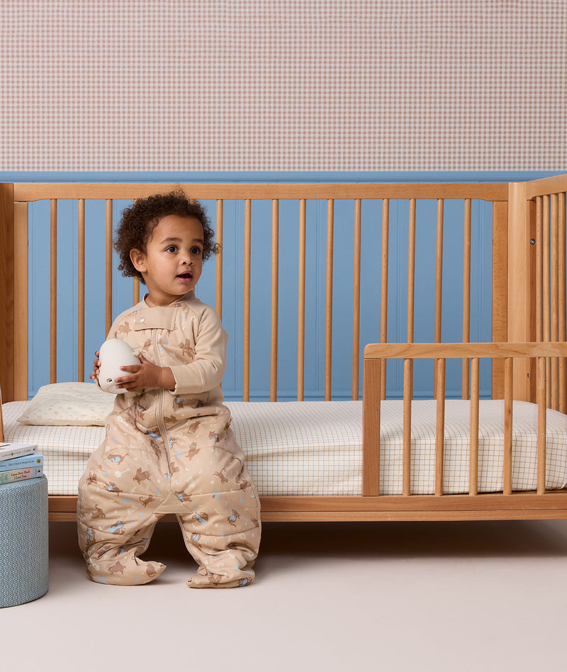 A toddler sits on the edge of a wooden toddler bed, wearing a beige patterned sleep suit bag with legs while holding a Blobby Bedtime Lamp.