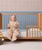 Smiling toddler wearing a beige patterned Sleep Suit bag, sitting on the edge of a wooden toddler bed.