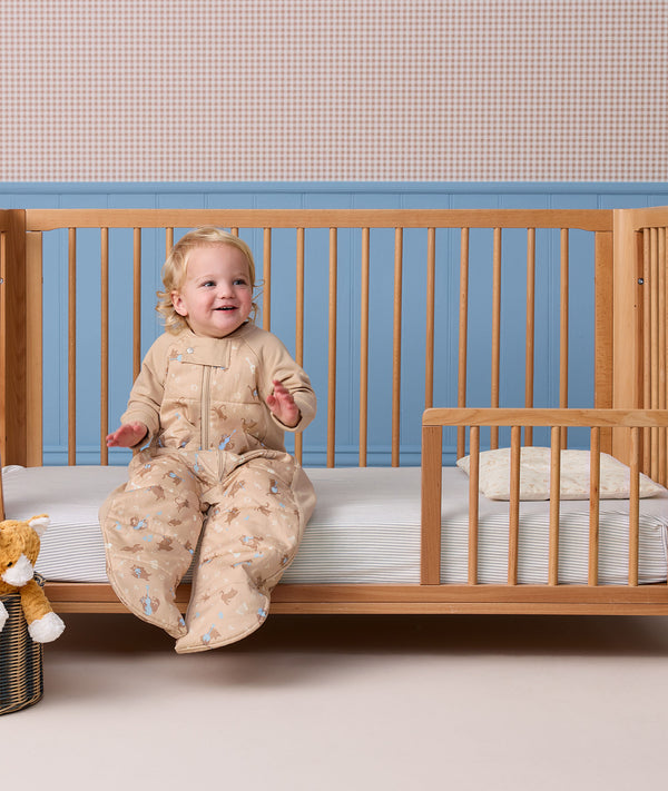 Smiling toddler wearing a beige patterned Sleep Suit bag, sitting on the edge of a wooden toddler bed.