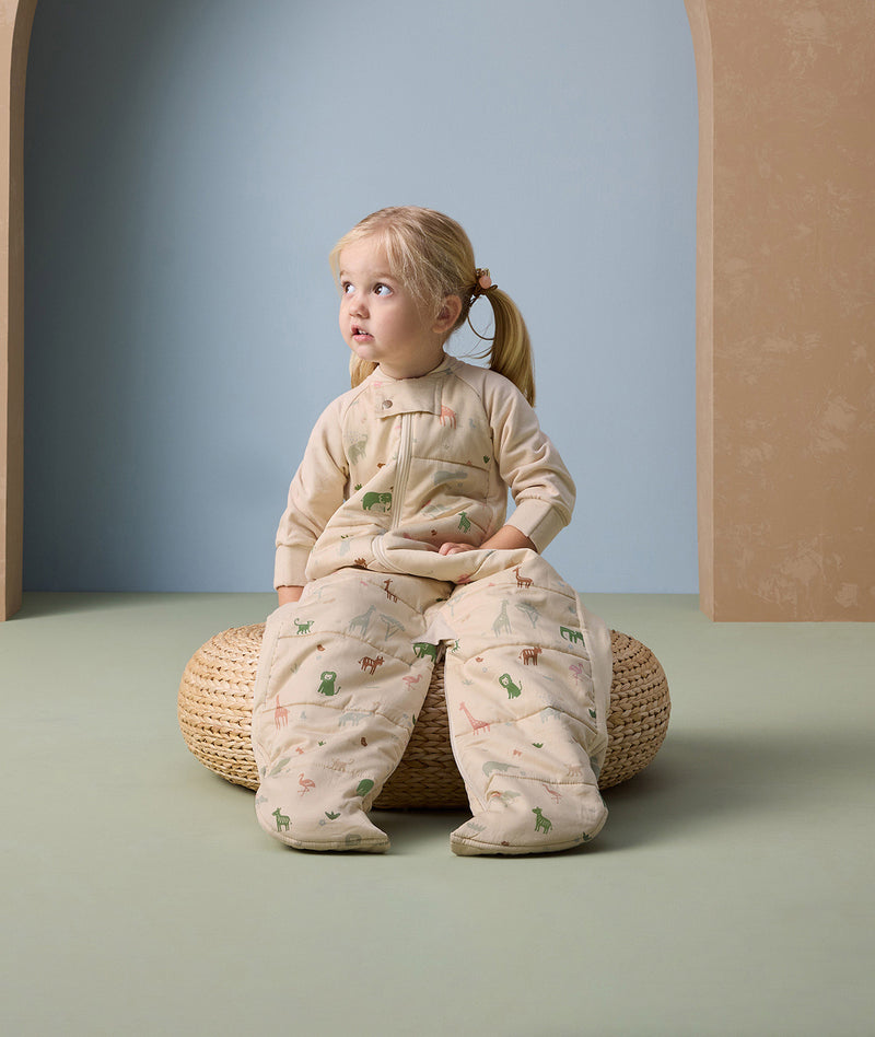 Toddler sitting on a woven cushion, wearing a beige sleep suit bag with legs, featuring a colorful animal print pattern including giraffes, elephants, and other safari animals.
