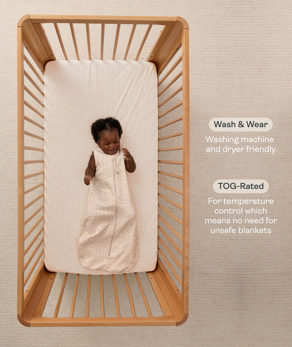 A happy baby lying inside a wooden cot, wearing a sleeveless ergoPouch Sleeping Bag 1.0 TOG with text highlighting 