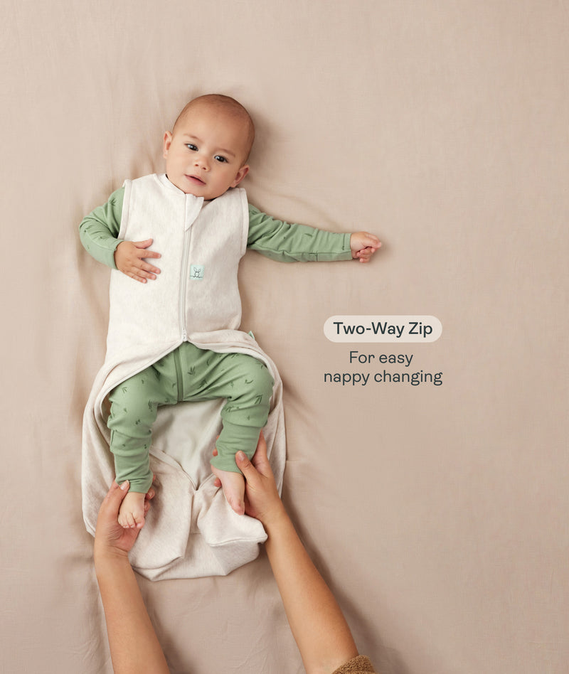 A baby lying down in a green long sleeve layer with an unzipped beige sleeping bag while adult hands hold their feet. Text highlighting 