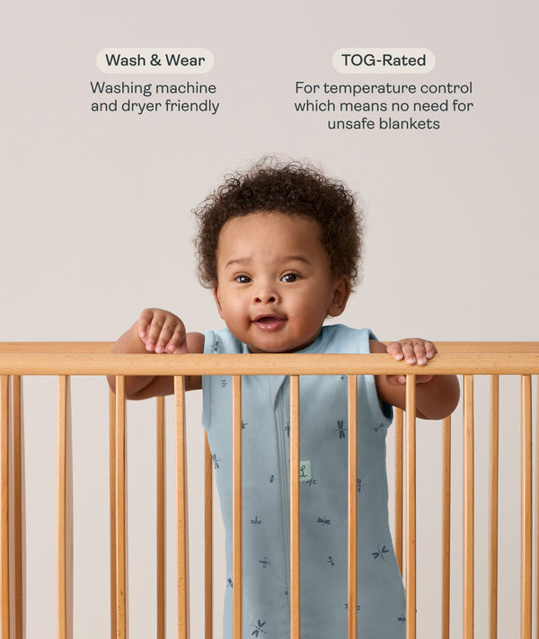 Infant wearing a sleeveless, light blue dragonfly-patterned sleeping bag stands in a wooden cot, holding onto the top rail with both hands. Above the baby, text features highlights for the product: Wash & Wear and TOG-Rated.