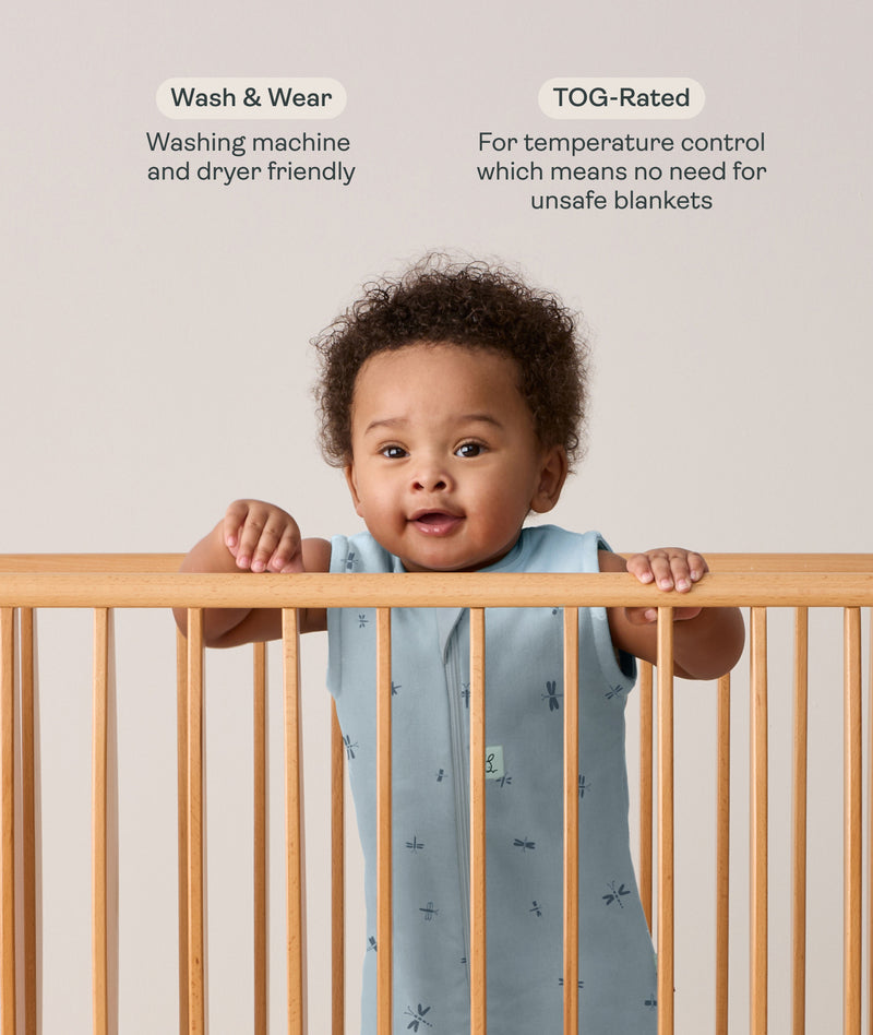 Infant wearing a sleeveless, light blue dragonfly-patterned sleeping bag stands in a wooden cot, holding onto the top rail with both hands. Above the baby, text features highlights for the product: Wash & Wear and TOG-Rated.