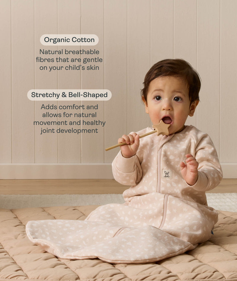 Infant sitting on a quilted mat while holding a wooden toy, wearing a long-sleeve patterned Sleeping Bag 1.0 TOG, with text highlighting organic cotton and stretchy and bell-shaped design.