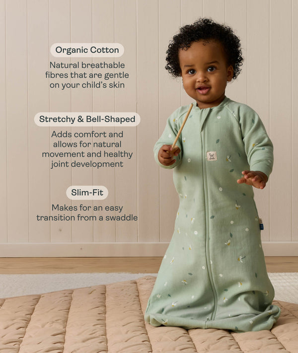 An infant wearing a long-sleeved sage green floral Sleeping Bag 1.0 TOG, standing on a padded beige mat with text highlighting 