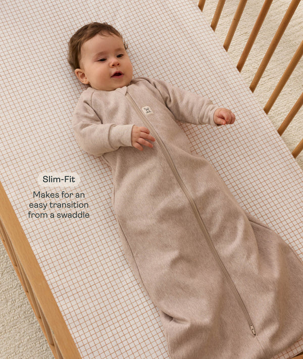 Infant lying in a wooden cot with a grid-patterned  sheet, wearing a long-sleeve Sleeping Bag 1.0 TOG with text highlighting its slim fit design for an easy transition form swaddling.