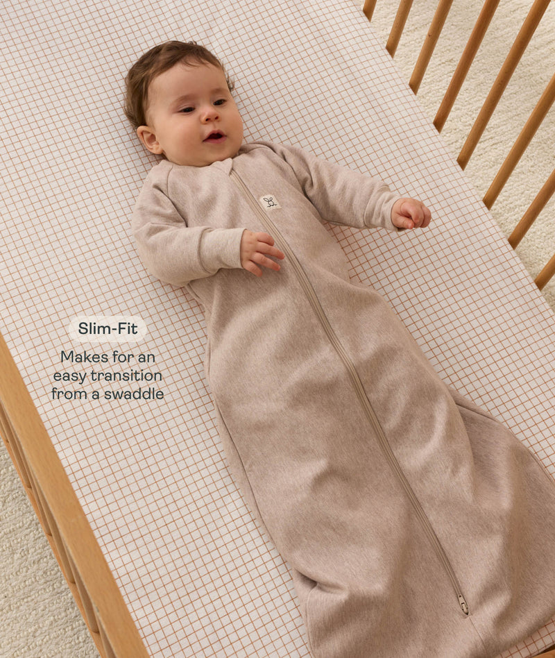 Infant lying in a wooden cot with a grid-patterned  sheet, wearing a long-sleeve Sleeping Bag 1.0 TOG with text highlighting its slim fit design for an easy transition form swaddling.