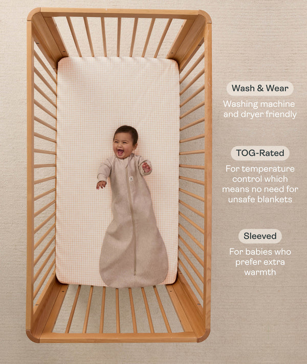 A happy baby lying in a wooden cot, wearing a sleeved Sleeping Bag 1.0 TOG Taupe Marle with text highlighting 