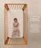 A happy baby lying in a wooden cot wearing a sleeveless Sleeping Bag 1.0 TOG Taupe Marle with text highlighting 