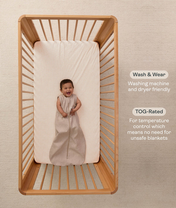 A happy baby lying in a wooden cot wearing a sleeveless Sleeping Bag 1.0 TOG Taupe Marle with text highlighting 