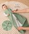 Baby lying on a padded mat while an adult adjusts the zipper of the baby's green sleeveless sleeping bag with text overlay highlighting two-way zip design for easy nappy changing and a close-up view of the dual zippers.