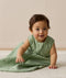 A baby wearing a sleeveless, leaf-patterned sleeping bag sits on a light rug, leaning forward with one hand resting on the floor.