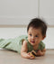A baby wearing a sleeveless, sage green leaf-patterned sleeping bag lies on their stomach on a wooden floor, holding a small wooden block and looking off to the side.