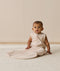 An infant wearing a sleeveless patterned Sleeping Bag 1.0 TOG, sitting upright on a white textured rug