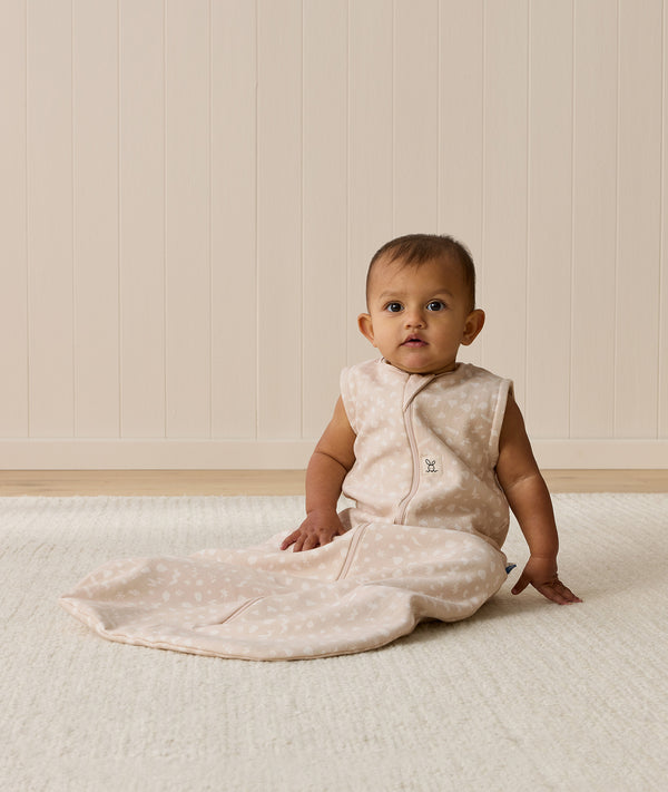 An infant wearing a sleeveless patterned Sleeping Bag 1.0 TOG, sitting upright on a white textured rug