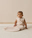 An infant wearing a sleeveless patterned Sleeping Bag 1.0 TOG, sitting upright on a white textured rug