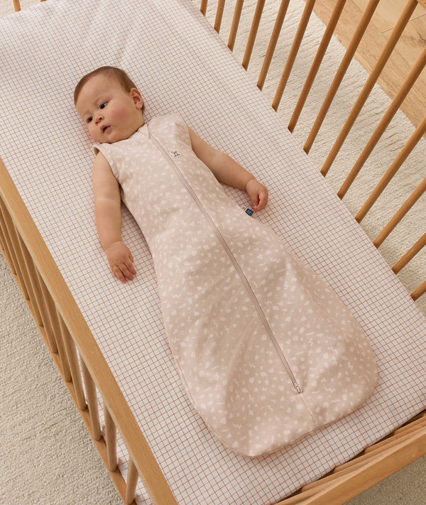 Infant wearing a beige patterned sleeveless Sleeping Bag 1.0 TOG, lying inside a wooden cot with a grid-patterned sheet