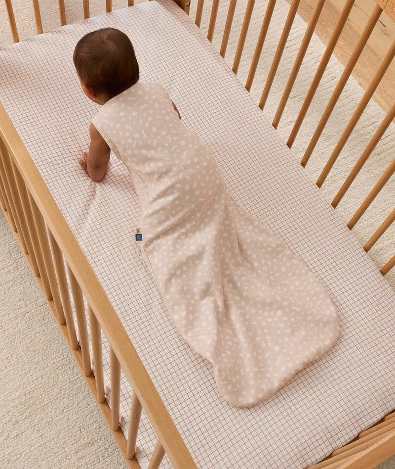 An overhead view of a baby wearing a pattern-printed sleeveless Sleeping Bag, crawling on their hands and knees inside a wooden cot with a grid sheet.