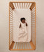 A smiling baby lying inside a wooden cot, wearing a sleeveless ergoPouch Sleeping Bag 1.0 TOG Coastal