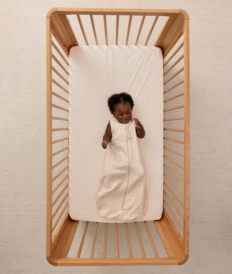 A smiling baby lying inside a wooden cot, wearing a sleeveless ergoPouch Sleeping Bag 1.0 TOG Coastal