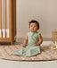 Infant sitting upright on a quilted mat, wearing a sleeveless sage green Sleeping Bag 1.0 TOG