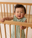A happy baby standing and holding onto the rail of a wooden cot, wearing a sleeveless Sleeping Bag 1.0 TOG with floral pattern.