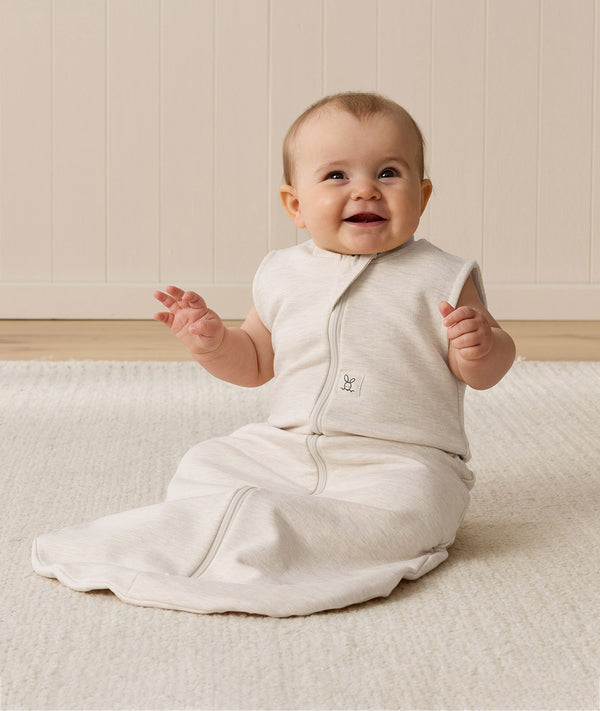 Smiling baby sitting on a white mat, wearing an ergoPouch Sleeping Bag 1.0 TOG Oatmeal Marle