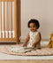 A smiling baby sitting on a beige quilted rug in a nursery while wearing a sleeveless, animal-patterned Sleeping Bag next to a wooden cot.