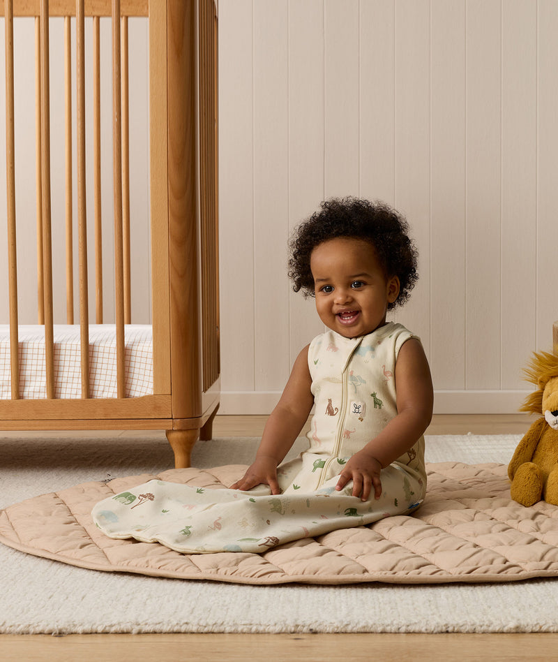 A smiling baby sitting on a beige quilted rug in a nursery while wearing a sleeveless, animal-patterned Sleeping Bag next to a wooden cot.