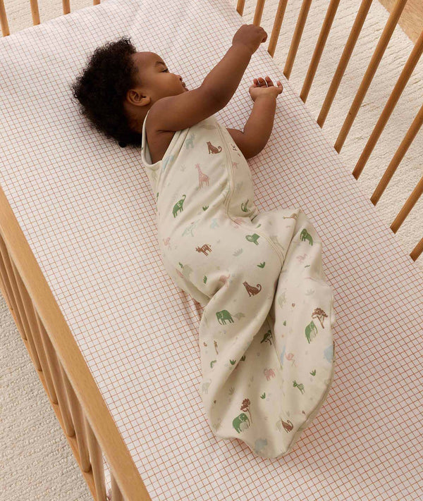 An infant wearing a cream safari-themed 1.0 TOG Sleeping Bag lying on its side in a wooden cot.