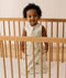 An infant standing inside a light wooden cot wearing an animal-patterned Sleeping Bag 1.0 TOG.