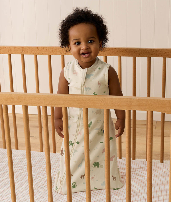 An infant standing inside a light wooden cot wearing an animal-patterned Sleeping Bag 1.0 TOG.