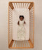 A happy baby lying inside a wooden cot, wearing a sleeveless, safari-themed Sleeping Bag.