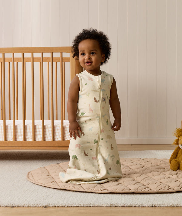 An infant standing upright on a beige quilted mat in a nursery while wearing an animal-patterned sleeveless Sleeping Bag next to a wooden cot.