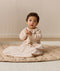 Infant sitting on a quilted mat while holding a wooden toy, wearing a long-sleeve patterned Sleeping Bag 1.0 TOG