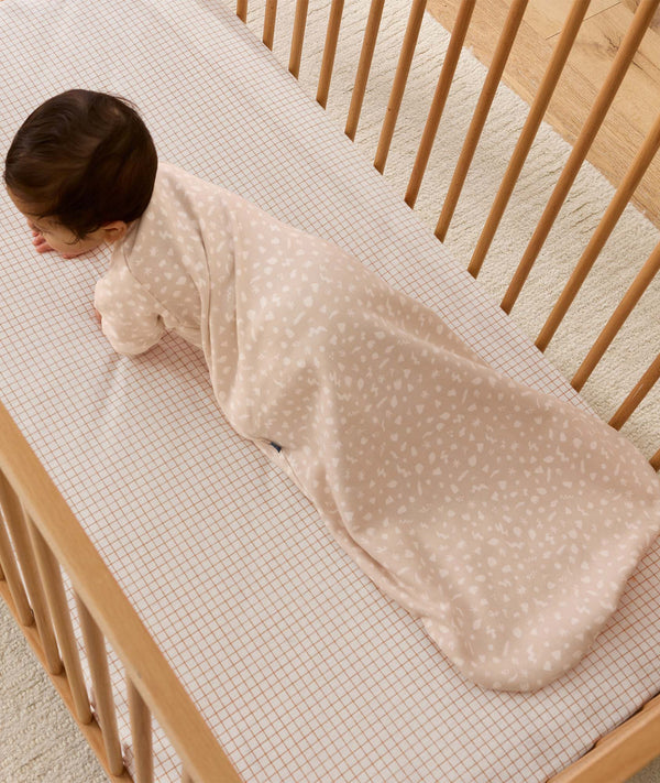 An overhead view of a baby wearing a pattern-printed long-sleeved Sleeping Bag, crawling on their hands and knees inside a wooden cot with a grid sheet.