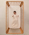 An overhead view on an infant lying in a wooden cot with a grid-patterned sheet, wearing a long sleeve Sleeping Bag 1.0 TOG
