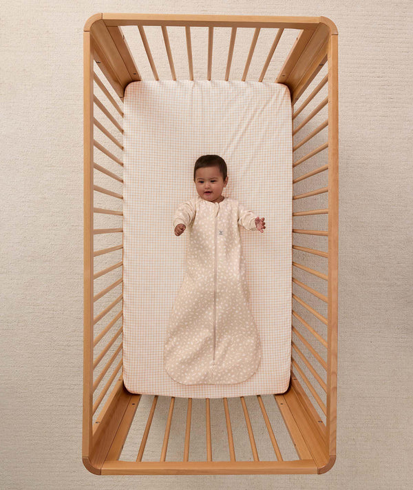 An overhead view on an infant lying in a wooden cot with a grid-patterned sheet, wearing a long sleeve Sleeping Bag 1.0 TOG
