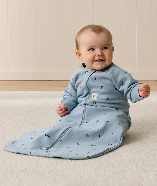 Smiling baby sitting on a white mat, wearing a blue sleeved Sleeping Bag 1.0 TOG with dragonflies pattern