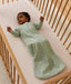 Baby lying on their back in a wooden cot, wearing a long-sleeved sage green floral Sleeping Bag 1.0 TOG over a grid-patterned sheet.