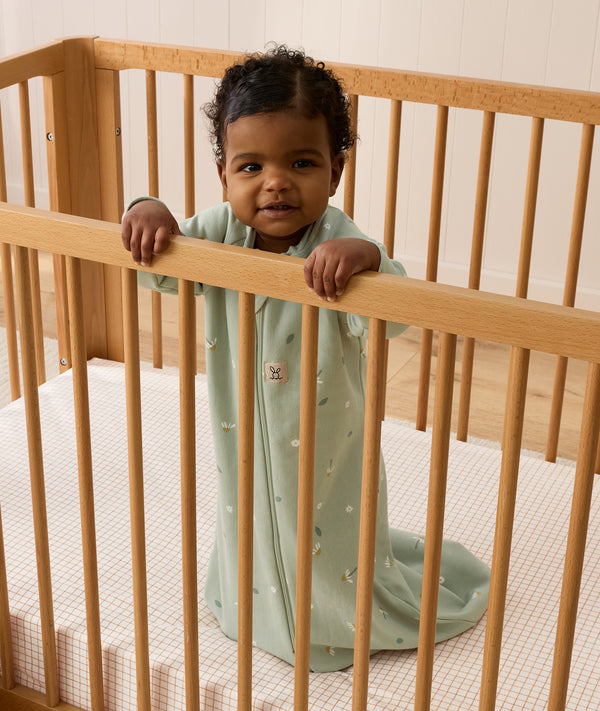 Infant wearing a sage green long-sleeve Sleeping Bag 1.0 TOG, standing inside a wooden cot