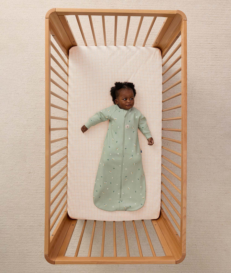 Infant lying inside a wooden cot with a grid-patterned sheet, wearing a long-sleeve Sleeping Bag 1.0 TOG