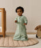 An infant wearing a long-sleeved sage green floral Sleeping Bag 1.0 TOG, standing on a padded beige mat next to a wooden cot.
