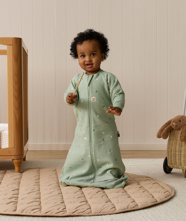 An infant wearing a long-sleeved sage green floral Sleeping Bag 1.0 TOG, standing on a padded beige mat next to a wooden cot.