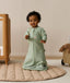 An infant wearing a long-sleeved sage green floral Sleeping Bag 1.0 TOG, standing on a padded beige mat next to a wooden cot.