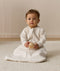 Infant wearing an ergoPouch Sleeping Bag 1.0 TOG Sleeved Oatmeal Marle, sitting on a white mat while holding a wooden toy.